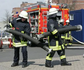Feuerwehrleute in Schutzkleidung tragen eine große Schlauchleitung vor einem Feuerwehrfahrzeug während einer Einsatzübung.